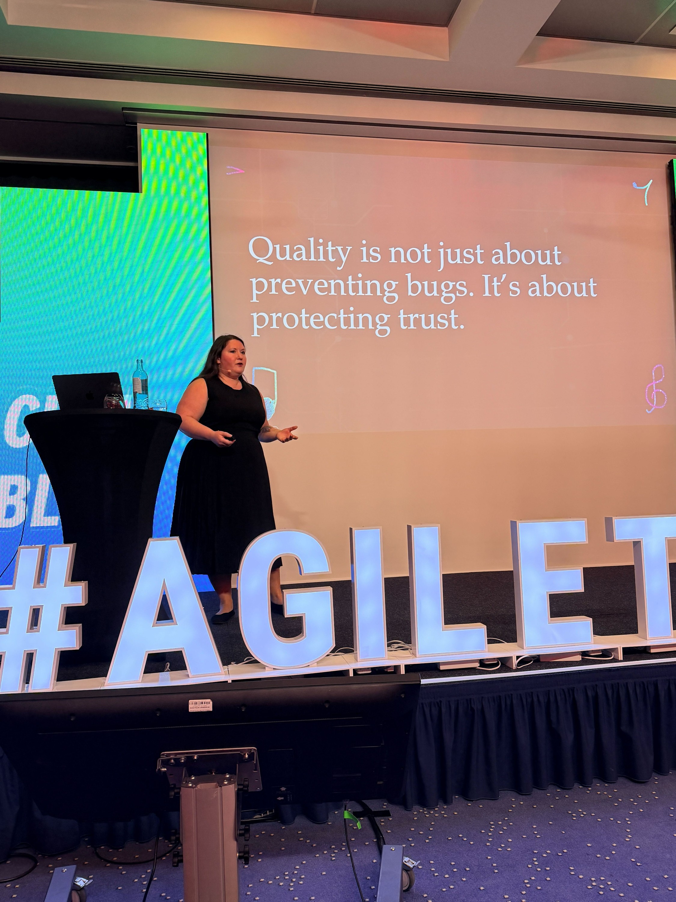 Rachel speaking on stage beside a slide reading “Quality is not just about preventing bugs. It’s about protecting trust.”